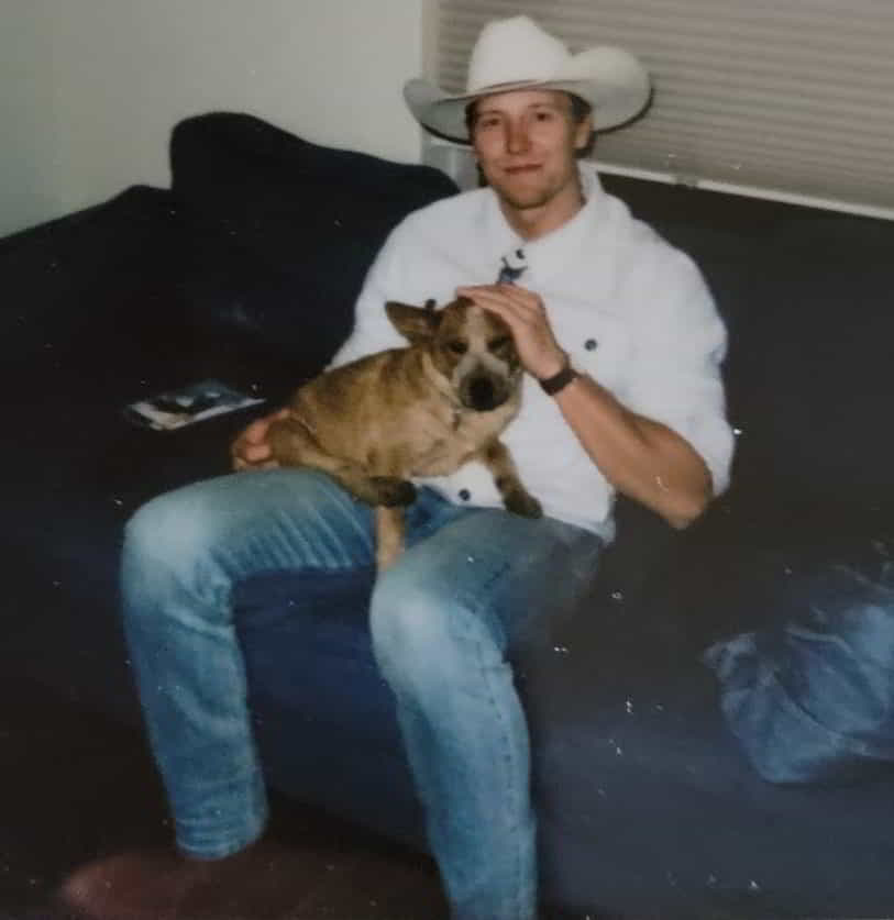 Polaroid picture of man wearing a cowboy hat and white button up shirt, sitting on a blue couch, petting a cute red heeler puppy. Both are looking at the camera.